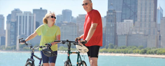 Two seniors smile at eachother as they stand beside heir bikes. They are along the lakeshore trail with the Chicago background in the background.